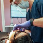 A dentist wearing a face shield and purple gloves uses a dental tool to carefully examine a patient's teeth.