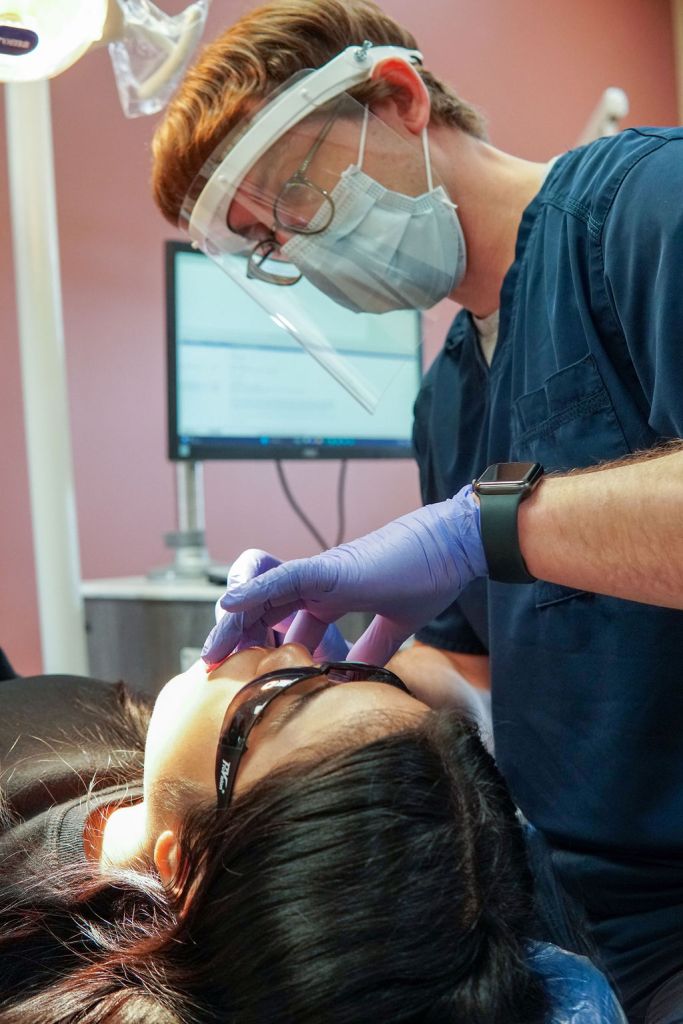 A dentist wearing a face shield and purple gloves uses a dental tool to carefully examine a patient's teeth.