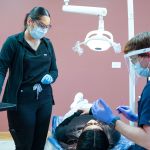A dental assistant and a dentist wearing protective face shields and masks prepare to treat a patient in a clinical setting.