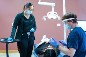 A dental assistant and a dentist wearing protective face shields and masks prepare to treat a patient in a clinical setting.