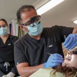 A male dentist wearing a mask, gloves, and magnifying loupes uses dental instruments and a suction tool on a patient's teeth.