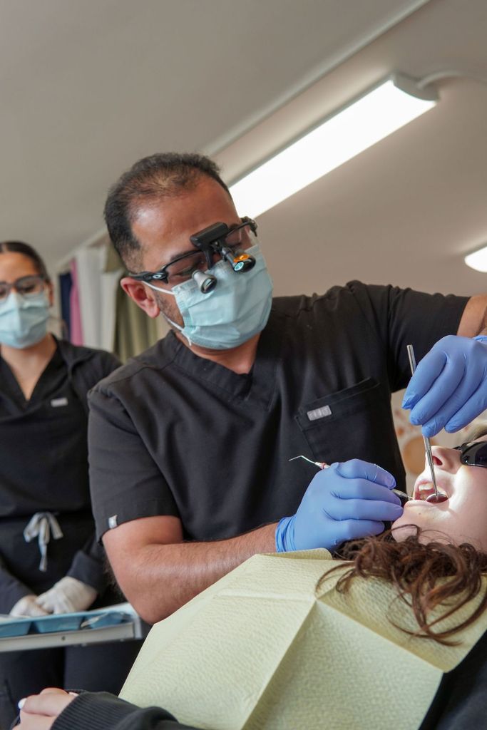 A male dentist wearing a mask, gloves, and magnifying loupes uses dental instruments and a suction tool on a patient's teeth.