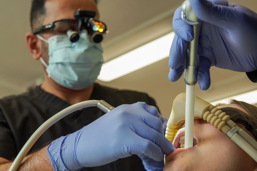 A male dentist wearing a mask, gloves, and magnifying loupes uses dental instruments and a suction tool on a patient's teeth.