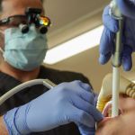 A male dentist wearing a mask, gloves, and magnifying loupes uses dental instruments and a suction tool on a patient's teeth.