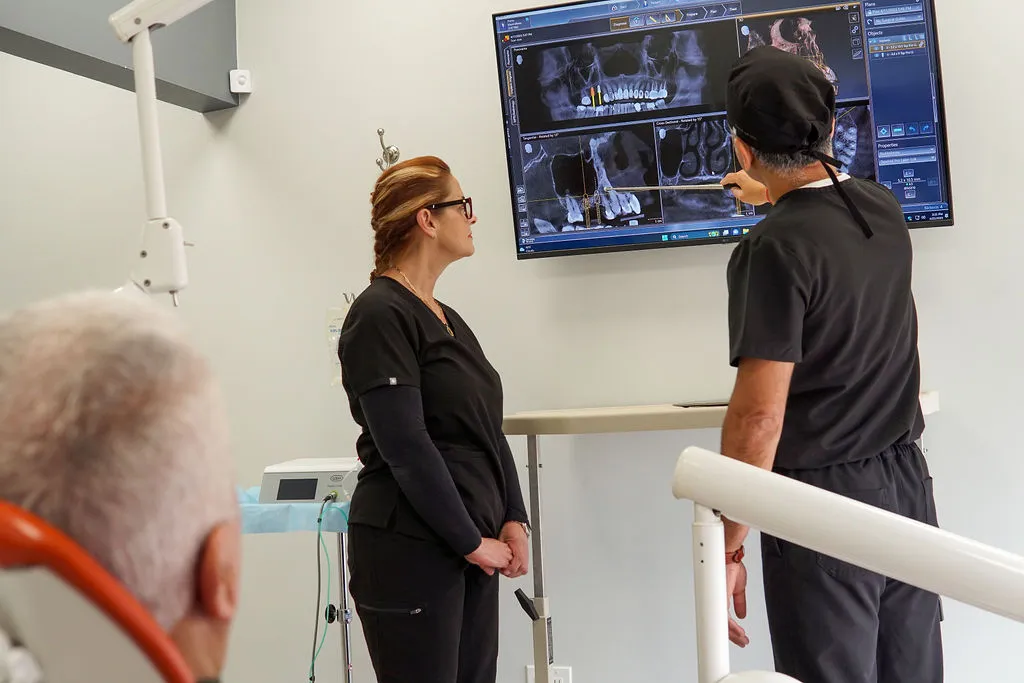 A female dental staff member in black scrubs stands with a male dentist in black scrubs, looking up at a wall-mounted monitor.