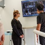 A female dental staff member in black scrubs stands with a male dentist in black scrubs, looking up at a wall-mounted monitor.