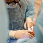 A gloved dental assistant's hands reach over a blue-draped surgical site to provide or take a small instrument for the lead surgeon.