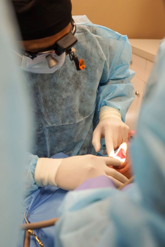A gloved dental assistant's hands reach over a blue-draped surgical site to provide or take a small instrument for the lead surgeon.