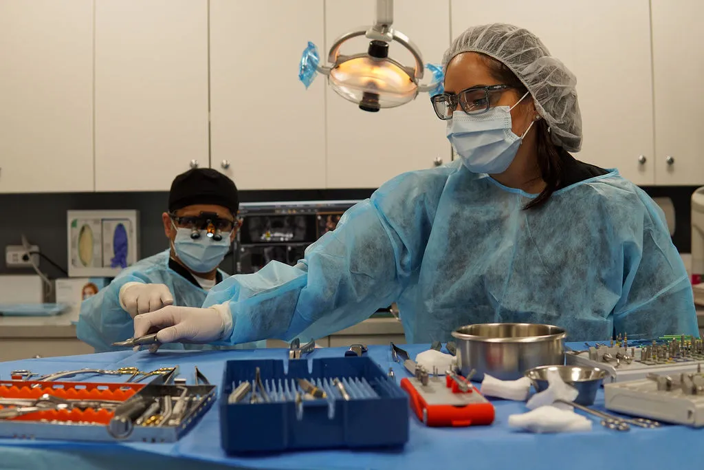Two dental assistants, wearing blue gowns, masks, and caps, prepare surgical instrument trays on a blue-draped table.