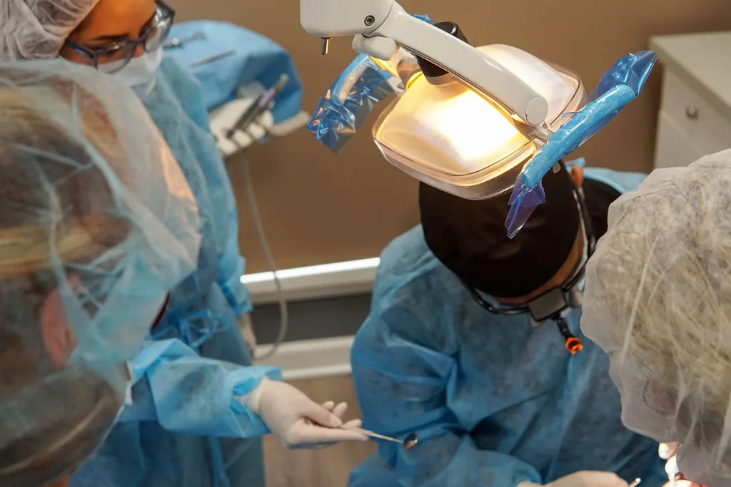 A high-angle view of a dental surgical team in blue sterile gowns and masks working on a patient under bright operatory lights.
