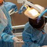A high-angle view of a dental surgical team in blue sterile gowns and masks working on a patient under bright operatory lights.