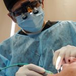 Close-up of a dental surgeon in a blue gown, cap, and mask, with magnifying loupes over their glasses, working on a patient.