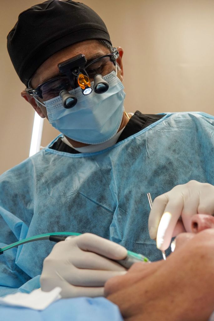 Close-up of a dental surgeon in a blue gown, cap, and mask, with magnifying loupes over their glasses, working on a patient.