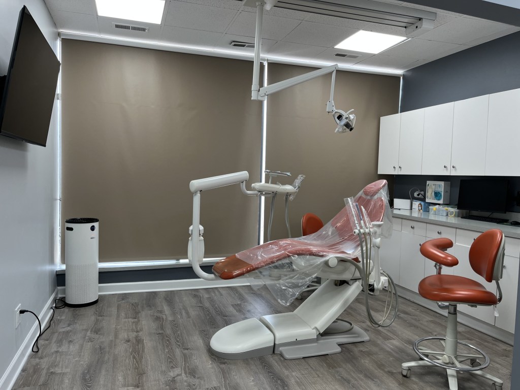 A contemporary dental operatory room equipped with a tan patient chair, overhead medical lighting, and cabinetry.