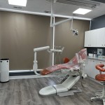 A contemporary dental operatory room equipped with a tan patient chair, overhead medical lighting, and cabinetry.