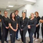 A professional team of seven dental staff members wearing matching black scrubs, standing together in a hallway and smiling.