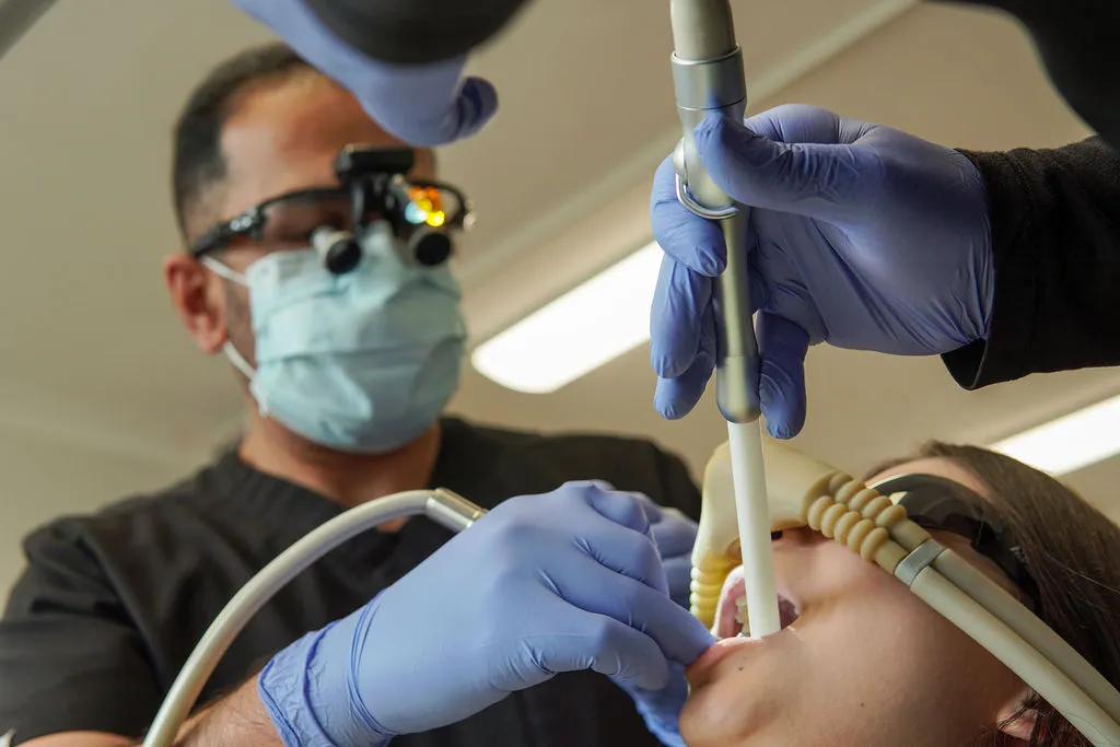 A male dentist wearing a mask, gloves, and magnifying loupes uses dental instruments and a suction tool on a patient's teeth.