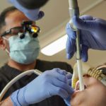 A male dentist wearing a mask, gloves, and magnifying loupes uses dental instruments and a suction tool on a patient's teeth.
