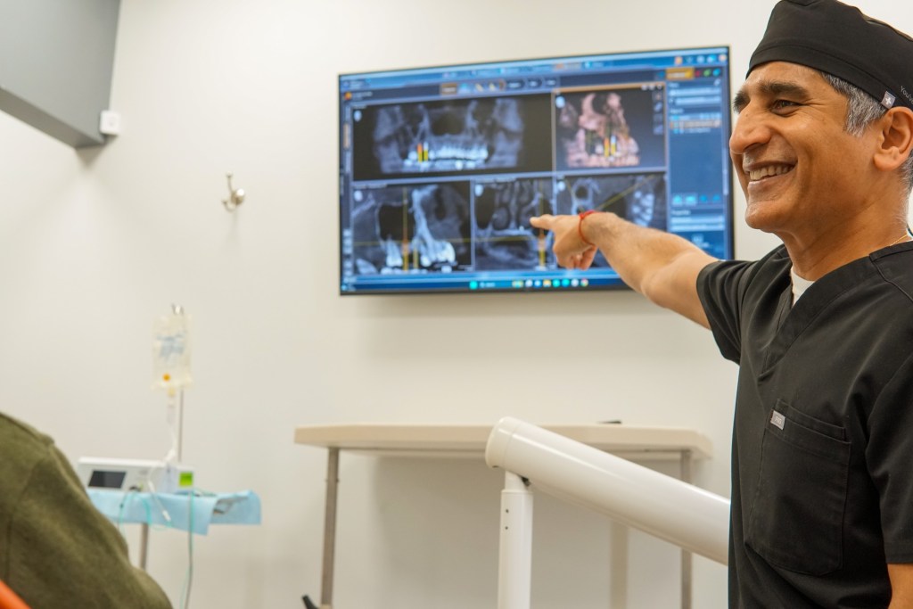 A male dentist in black scrubs points towards a large wall-mounted monitor showing multiple screens, in a dental office.