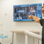 A male dentist in black scrubs points towards a large wall-mounted monitor showing multiple screens, in a dental office.