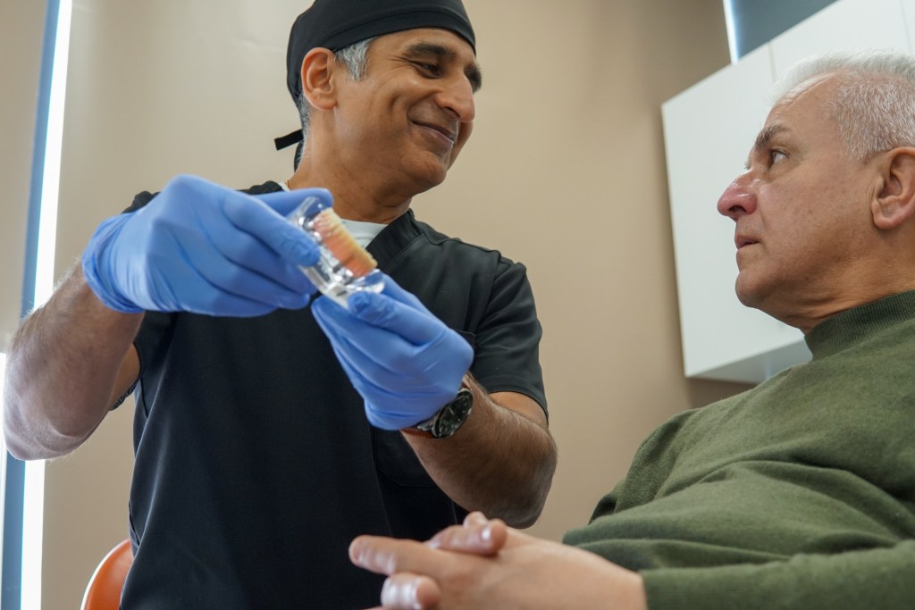 A male dentist in black scrubs and blue gloves holds a small dental model while explaining a procedure to an older male patient.