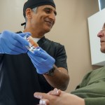 A male dentist in black scrubs and blue gloves holds a small dental model while explaining a procedure to an older male patient.