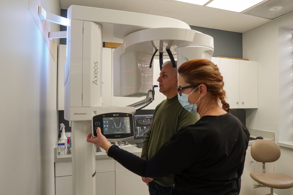 A dental professional assisting an older male patient as he prepares to take a panoramic dental X-ray with a large white digital imaging machine.