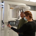 A dental professional assisting an older male patient as he prepares to take a panoramic dental X-ray with a large white digital imaging machine.