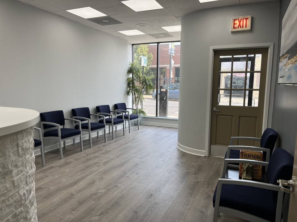 Interior view of a bright dental office waiting area with blue chairs, wood-style flooring, and a glass entrance door.