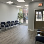 Interior view of a bright dental office waiting area with blue chairs, wood-style flooring, and a glass entrance door.
