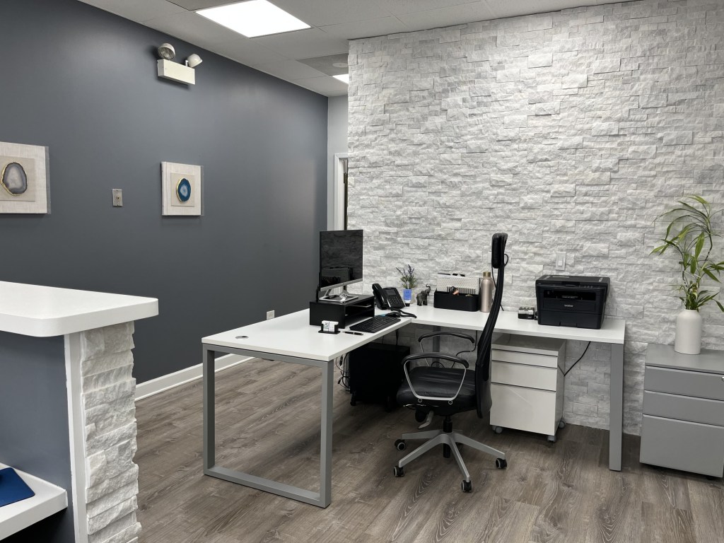 A modern dental consultation room featuring a white desk with a computer monitor and a textured white accent wall.